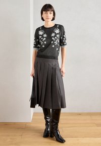 Μαύρος σπαστός πουλόβερ με λουλουδάτη κεντημένη διακόσμηση, σε συνδυασμό με μια πλυμένη γκρι φούστα και μαύρες μπότες μέχρι το γόνατο. Μοντέλο κρατώντας μια μαύρη τσάντα.