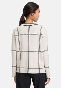 Light beige sweater with black grid pattern, knit fabric, open collar, and long sleeves, featuring a straight hem and minimalistic design.
