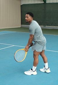Un jugador de tenis con un atuendo atlético de color gris claro se prepara para sacar. Sosteniendo una raqueta amarilla, lleva zapatillas blancas con detalles en negro, en una pista azul.