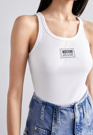 Femme portant un débardeur blanc côtelé avec un patch logo "Moschino Jeans", associé à un jean en denim bleu taille haute.