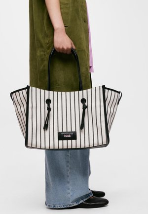 Osoba w zielonej kamizelce i dżinsach trzyma dużą białą torbę typu tote z czarnymi pionowymi paskami i czarnymi uchwytami.