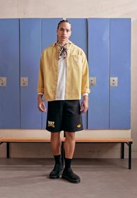 Yellow oversized shirt, white t-shirt, black shorts with yellow print, black socks, and clogs. Background features blue lockers.