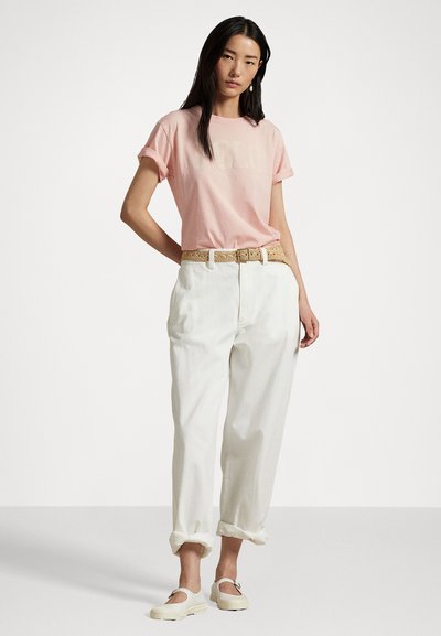 Lys rosa t-skjorte med rullet ermer og et subtilt grafisk motiv, kombinert med løse hvite bukser og et beige flettet belte. Hvite slip-on sko fullfører antrekket.