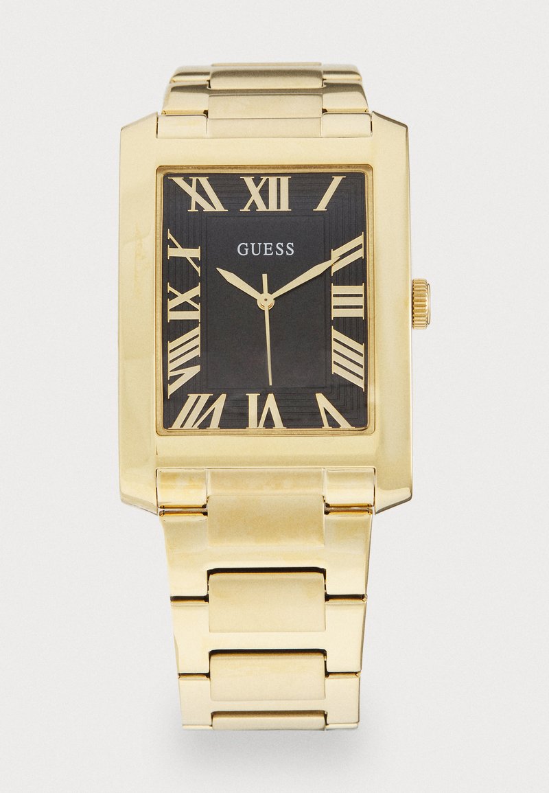 Guldfarvet rektangulært ur med sort urskive, romerske tal, og det mærkede "GUESS" logo. Metalarmbåndet har polerede led og en skinnende finish.