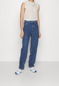 Top blanco sin mangas, ligero y con detalles en relieve, jeans de mezclilla de tiro alto azules y zapatillas deportivas grises con detalles azules.