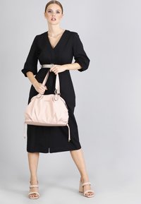 Różowa skórzana torebka o marszczonym wzorze, z podwójnymi uchwytami i złotymi elementami. Nosi ją w połączeniu z czarną sukienką i beżowymi szpilkami.