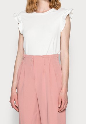 Kobieta w białym topie bez rękawów z falbanami na ramionach oraz różowych, plisowanych spodniach z wysokim stanem.