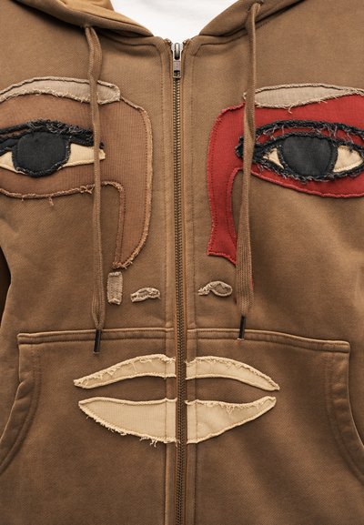 Καφέ φούτερ με φερμουάρ, με κεντημένο σχέδιο προσώπου σε πολλά χρώματα—μαύρο, κόκκινο και μπεζ. Υφή υφάσματος με ξεφτισμένα άκρα.