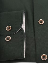 Donkergroen shirt met een herringbone-textuur, contrasterende witte voering en bruine knopen met vier gaten. Close-up van de zak en de kraag.