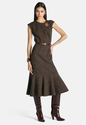 Femme portant une robe marron texturée sans manches avec un bas évasé, ceinture marron, bottes hautes en cuir marron, ainsi qu'une broche et un bracelet décoratifs.