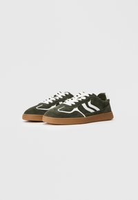 Ein Paar dunkelgrüne Wildledersneaker mit weißen Schnürsenkeln, weißen Seitenstreifen und braunen Gummisohlen auf einem schlichten hellen Hintergrund.