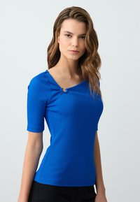 Top blu corto a maniche con scollo a barchetta e dettaglio decorativo a loop sul davanti. Realizzato in un tessuto liscio e elasticizzato.