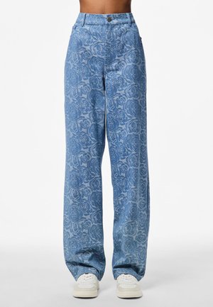 Jeansy z wysokim stanem i szerokimi nogawkami w kolorze niebieskim z różanym wzorem, zapinane z przodu na guziki, noszone z białymi trampkami i odsłoniętym, krótkim brzuchem.