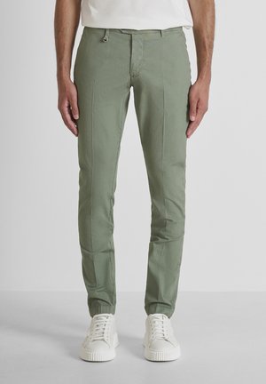 Homme portant un pantalon ajusté vert olive et des baskets blanches, debout devant un fond gris clair uni.