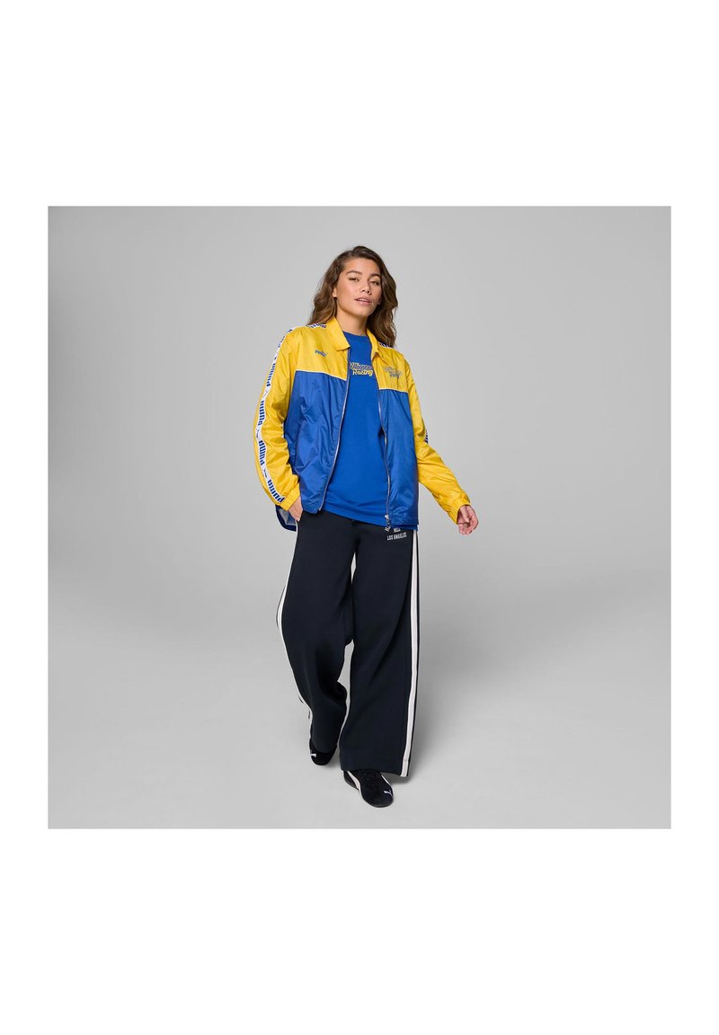 Niebieska i żółta kurtka z zamkiem z przodu, w połączeniu z niebieską koszulą oraz luźnymi czarnymi spodniami z białymi paskami po bokach.