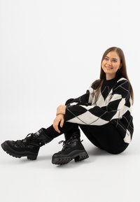 Pull en argyle noir et blanc avec un col montant, associé à des bottes de cheville isolées noires avec des semelles épaisses et des panneaux texturés.