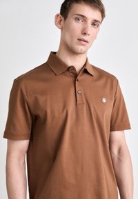 Barna, sima pamutból készült póló, galléros nyakkal, két gombbal és egy kis logóval a bal mellen.