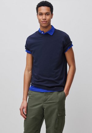 Młody mężczyzna w nawarstwionych granatowo-niebieskich koszulach z podwiniętymi rękawami, oliwkowo-zielonych spodniach typu cargo, pozujący z jedną ręką w kieszeni.