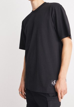 Person trägt ein schlichtes schwarzes T-Shirt mit kurzen Ärmeln und einem kleinen weißen "CK" Logo-Patch nahe dem unteren Saum auf der linken Seite.
