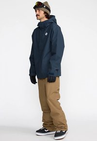 Marineblauwe waterdichte jas met capuchon, beige sneeuwbroek, zwarte handschoenen en zwarte schoenen met witte accenten; de jas heeft een logo.