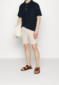 Man i svart kortärmad polo, beige shorts, svarta och orange sandaler, håller en vikt vit väska mot en jämn vit bakgrund.