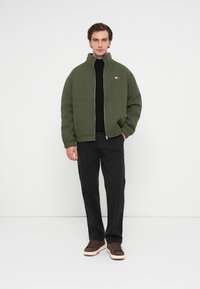 Πράσινο φλις μπόμπερ μπουφάν με ψηλό γιακά και κλείσιμο με φερμουάρ, συνδυασμένο με μαύρα παντελόνια και καφέ αθλητικά παπούτσια. Λογότυπο στο στήθος.