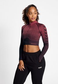 Fitted, cropped Langarmshirt in Bordeaux, mit einem halben Reißverschluss und kontrastierenden schwarzen Chevron-Mustern. Kombiniert mit lockeren schwarzen Hosen.