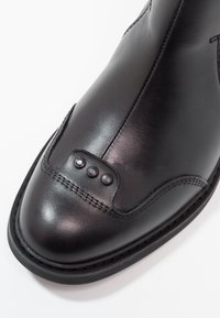 Chaussure en cuir noir en gros plan montrant une surface lisse, des coutures détaillées et trois clous décoratifs noirs près de la zone des orteils.
