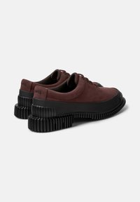 Scarpe in suede borgogna con suole in gomma nera, dotate di punte arrotondate e lacci, esposte da un angolo leggero per mostrare la vista posteriore.