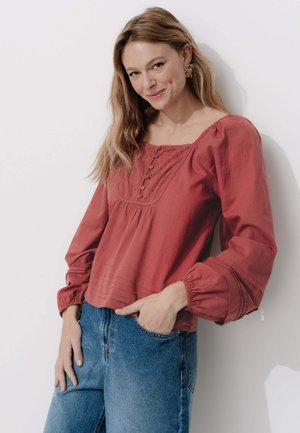 Blouse couleur rouille avec un décolleté carré, boutons décoratifs à l'avant, détails plissés et manches ballon. Associée à un jean en denim bleu.