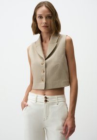 Beige ärmellose Cropped-Weste aus strukturiertem Stoff mit zwei großen Knöpfen und einem Reverskragen. Kombiniert mit hochtaillierten weißen Hosen.