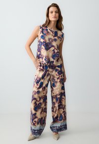 Ärmelloser Jumpsuit mit Blumenmuster, der eine beige Grundfarbe und aqua- und violetten Paisleymustern aufweist, weit geschnittene Hosenbeine und seitliche Taschen hat.
