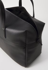 Sac en cuir noir avec un extérieur texturé, des anses lisses et une fermeture zippée supérieure. L'intérieur est doublé et spacieux, conçu pour la polyvalence.