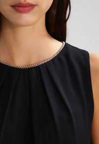 Robe noire sans manches avec un corsage froncé et un détail de d écolleté orné de perles argentées, mettant en valeur une texture de tissu lisse.