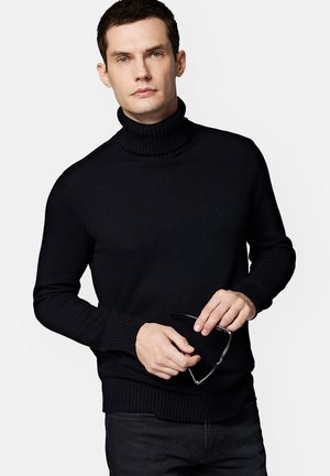 Czarny sweter z golfem o prążkowanej fakturze i ściągaczach. Trzymane okulary w dłoniach. Prosty design z dyskretnym logo z przodu.