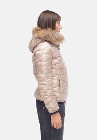 Giacca puffer beige con cappuccio bordato in pelliccia, motivo trapuntato e due tasche laterali. Il tessuto ha una texture liscia e lucida.