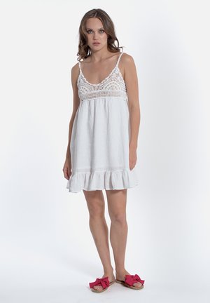 Robe en lin blanche avec un corsage en crochet et un ourlet froncé en plusieurs niveaux. Dotée de fines bretelles et d'une coupe décontractée. À porter avec des sandales à nœud rose.