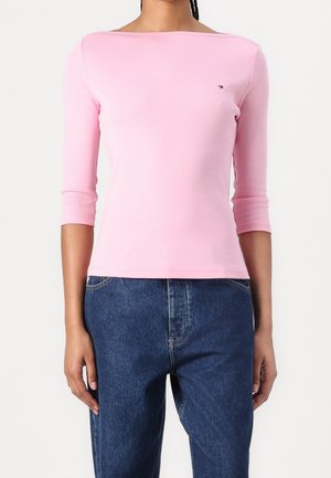 Kvinde iført en tætsiddende lys lyserød bådudskæring top med trekvartlange ærmer og højtaljede blå denimjeans.