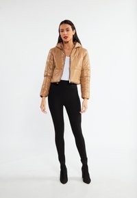 Sandfarvet puffjakke med hætte, cropped design, skinnende overflade, frontlynlås og sorte højtaljede leggings. Sorte ankelstøvler.