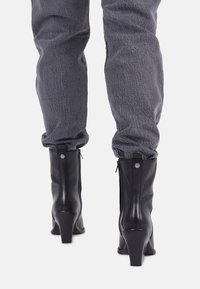 Czarne skórzane botki za kostkę o spiczastym nosku i słupkowym obcasie, z bocznym zamkiem błyskawicznym i metalowymi akcentami, zestawione z szarymi, teksturowanymi spodniami.