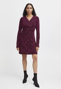 Kapeita, long-sleeve dress in deep magenta with diamond pattern. Soft fabric, V-neckline, and mid-thigh length. Worn with sheer black heels.