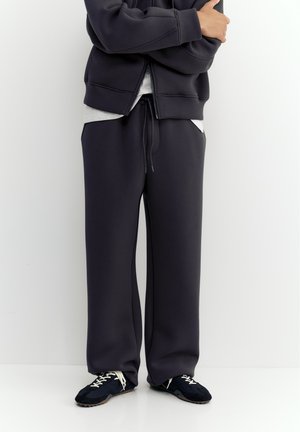 Personne portant un pantalon de survêtement noir avec cordon de serrage, des baskets noires avec des lacets blancs, un sweat à capuche zippé noir et une chemise blanche, debout contre un mur blanc.