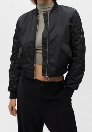 Femme portant une veste bomber noire avec une fermeture éclair argentée, un col roulé court olive et un pantalon noir taille haute sur un fond uni.