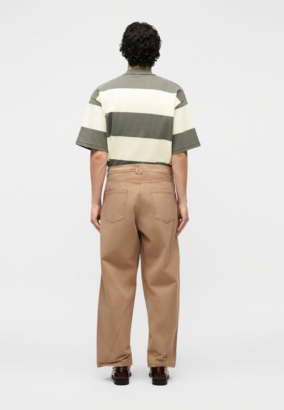 Άνδρας με την πλάτη γυρισμένη φοράει κοντομάνικο πουκάμισο με οριζόντιες ρίγες σε ελιά και εκρού χρώμα, φαρδιά καφέ παντελόνια και σκούρα καφέ παπούτσια σε απλό φόντο.