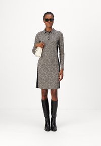 Schwarz-beige Leopardenmuster Kleid mit langen Ärmeln, einem Kragen und seitlichen Schlitzen. Kombiniert mit kniebedeckenden, glänzenden schwarzen Stiefeln und einer weißen Tasche.