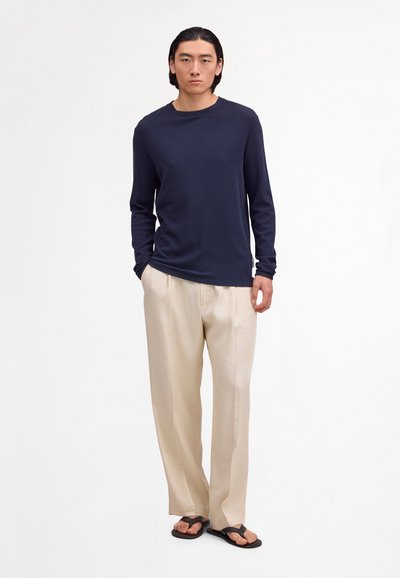 Muž s rovnými čiernymi vlasmi, oblečený v tmavomodrej dlhorukávovej košeli, béžových širokých nohaviciach a čiernych sandáloch, stojaci pred bielym pozadím.