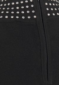 Svart stickad plagg med en central dragkedja, prydd med små, glänsande, cirkulära nitar längs den övre delen för ökad textur.