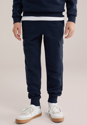 Isik kannab tumesiniseid dressipükse küljetaskutega, valgeid sokke, valgeid tossusid beežide detailidega ja tumesinist dressipluusi.