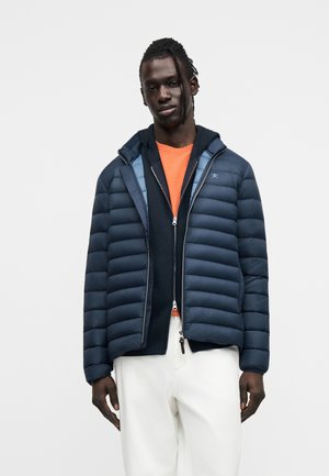 Man wearing a navy puffer jacket over a black zip-up hoodie and orange shirt, paired with white pants against a plain white background.