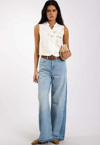 Top blanco sin mangas con textura floral, combinado con jeans de pierna ancha azul claro y un cinturón marrón. Sosteniendo un bolso beige de punto.